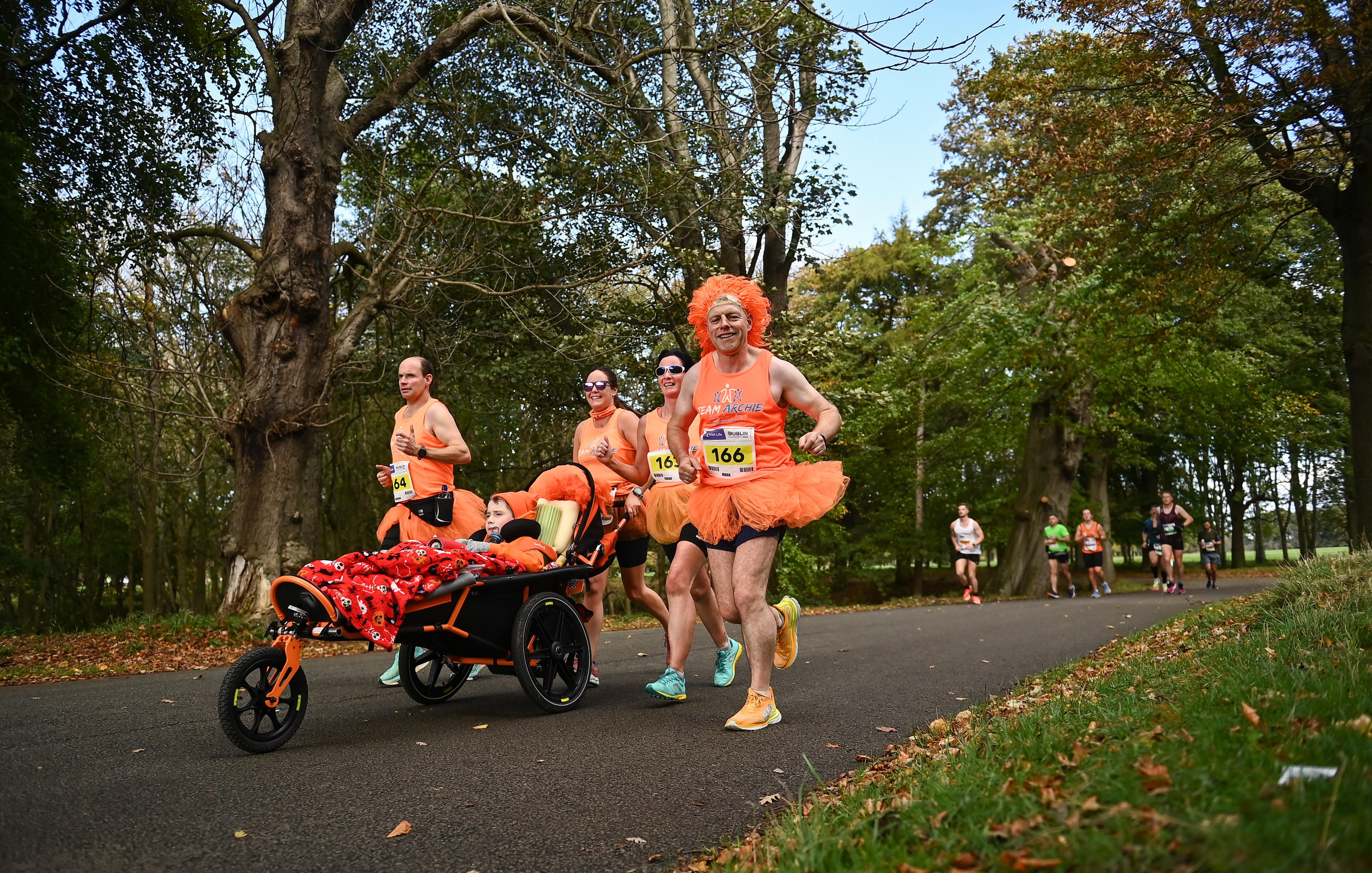 Four people running in orange tutus and wigs, with a child in a running wheelchair.